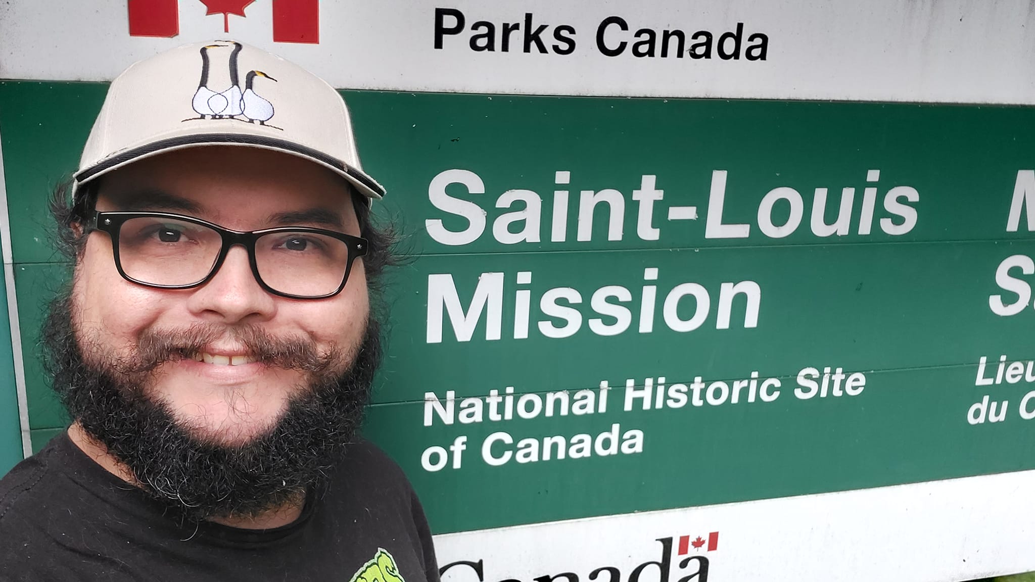 A man with short hair and bushy facial hair stands in front of a green and white sign that reads "Parks Canada, Saint-Louis Mission, National Historic Site of Canada"1